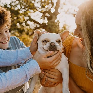 A sunlit photo of a boy and a young woman making a fuss of a white French Bulldog held between them outdoors.