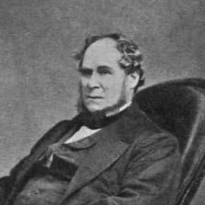 A black-and-white Victorian portrait photograph of a seated man with mutton-chop sideburns in a dark suit.